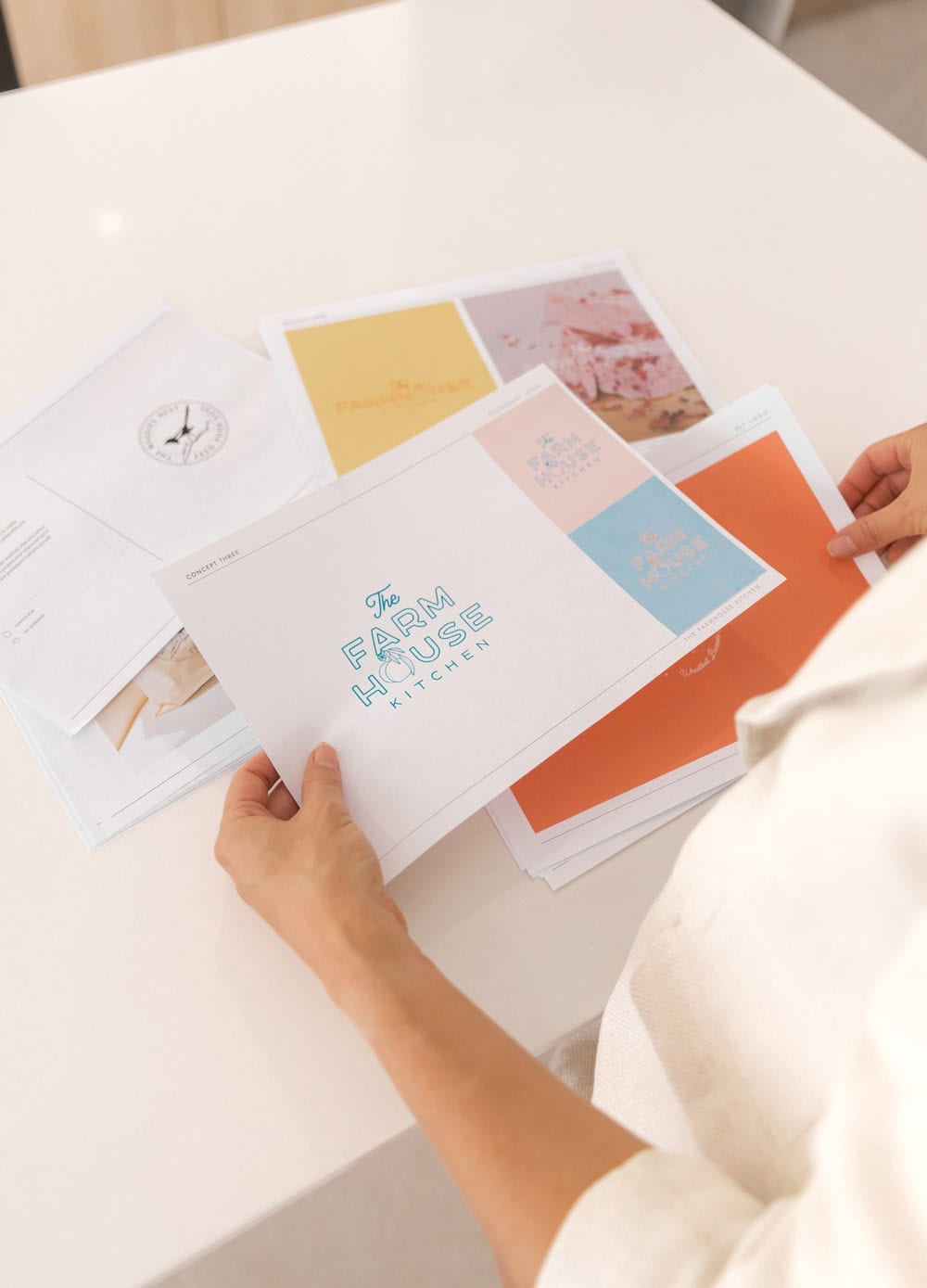 Sally J holding a printed sheet with "The Farm House Kitchen" logo among several branding design Perth drafts spread on a white table.