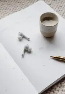 Open planner with a pair of wireless earbuds, a ceramic cup of coffee, and a gold pen placed on top, set on a textured light-colored surface.